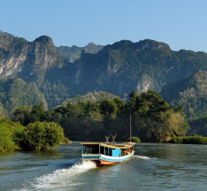 Langsames Holzboot fährt auf einem breiten Fluss, umgeben von dicht bewaldeten Bergen im goldenen Abendlicht.