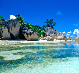 Traumstrand auf den Seychellen mit markanten Granitfelsen, kristallklarem Wasser und üppiger tropischer Vegetation unter strahlend blauem Himmel.