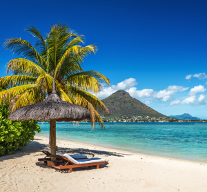 Idyllischer Sandstrand auf Mauritius mit Liege unter einer Palmen-Schilf-Sonnenschirm, türkisblauem Meer und Blick auf einen markanten grünen Berg unter strahlend blauem Himmel.