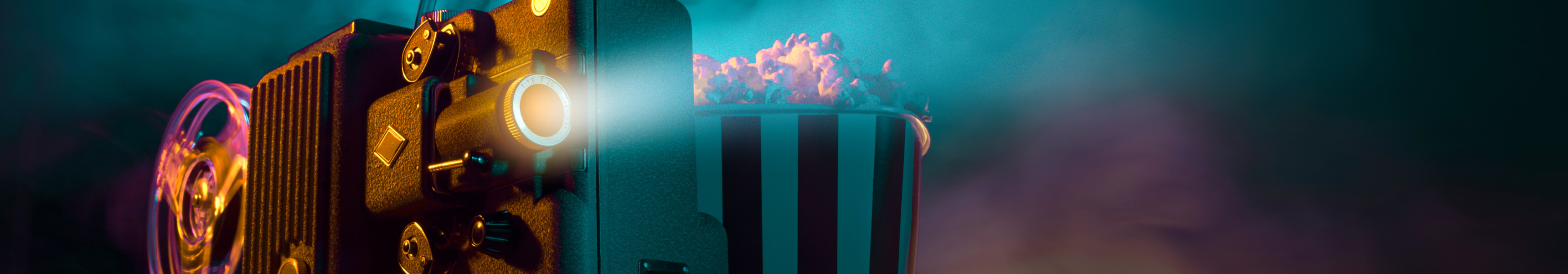 Alter Filmprojektor mit laufender Filmrolle, daneben eine gestreifte Popcorntüte, im Hintergrund bunte Licht- und Raucheffekte.