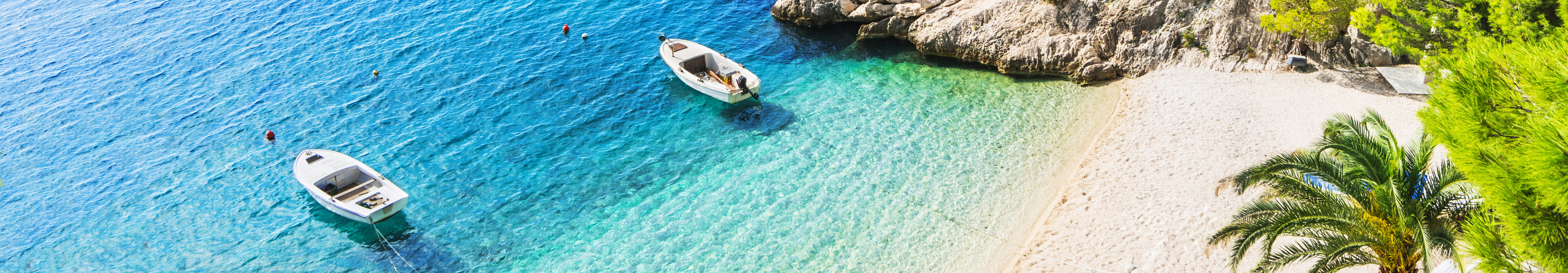 Idyllischer Strand in Brela an der Makarska Riviera in Kroatien mit türkisfarbenem Wasser, weißen Kieseln, kleinen Booten und Pinienbäumen am Ufer.