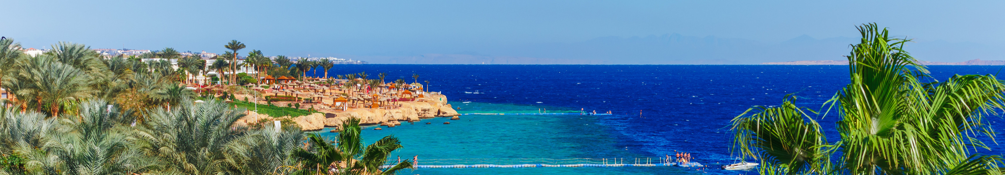Tropische Strandlandschaft mit türkisfarbenem Wasser, vielen Palmen im Vordergrund und Badegästen im Meer, im Hintergrund felsige Küste und blaues Meer bis zum Horizont.