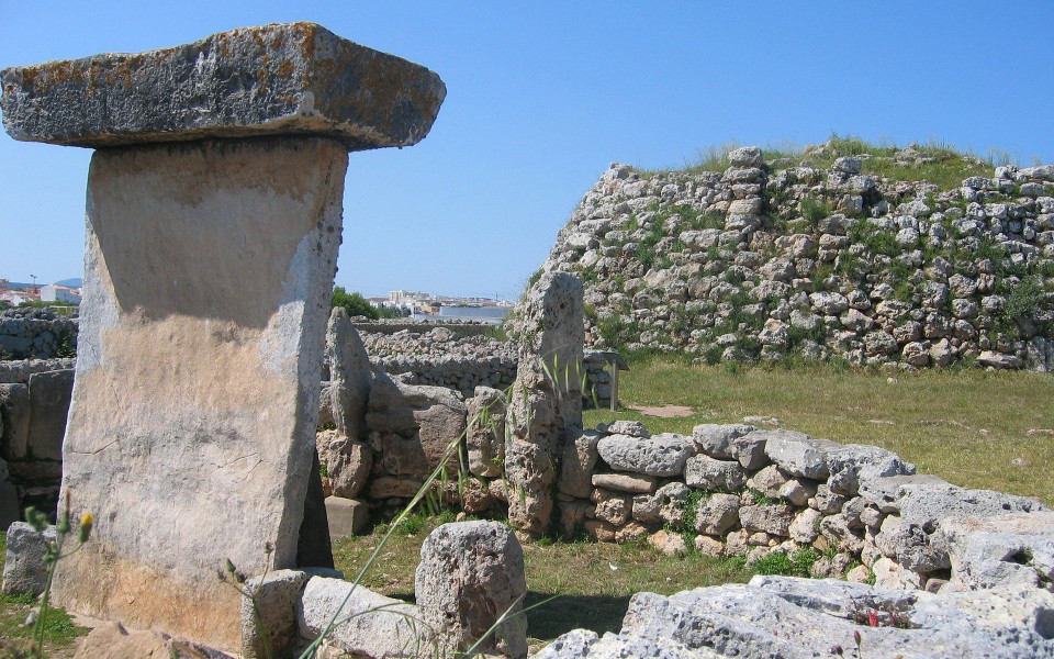 Prähistorische Ruinen der Talayot-Kultur auf Menorca mit großen Steinen und Mauern, umgeben von einer grünen Landschaft und blauen Himmel.