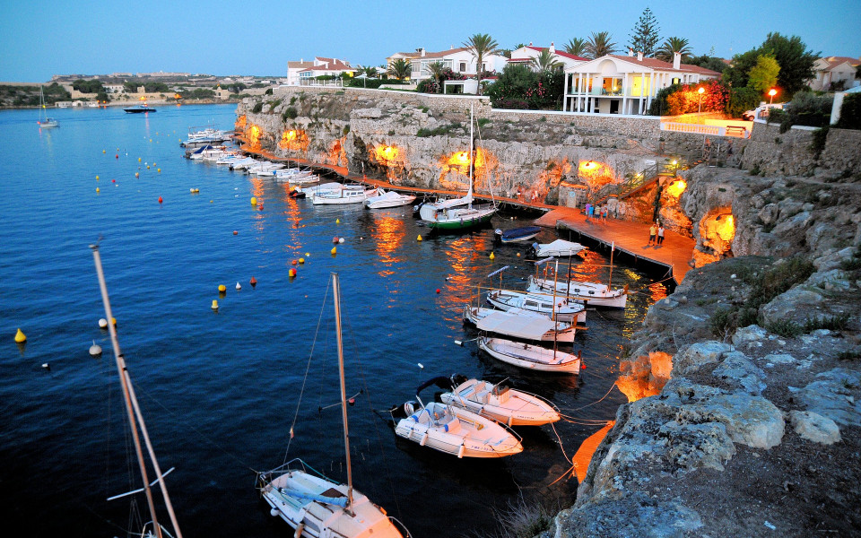 Hafen in Menorca mit Boote am Abend, umgeben von beleuchteten Felsen und weißen Häusern. Der Himmel ist leicht bewölkt und die Atmosphäre ist ruhig.