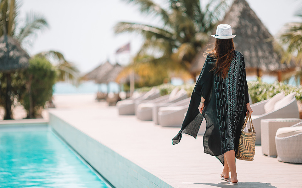 Frau im Sommerkleid mit Strohhut spaziert barfuß am Pool eines tropischen Resorts.