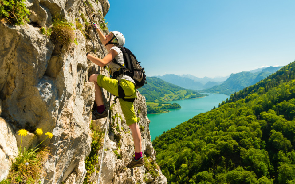 Kletterin sichert sich am Felsen mit Klettersteigset, im Hintergrund liegt der türkisfarbene Attersee im Salzkammergut.