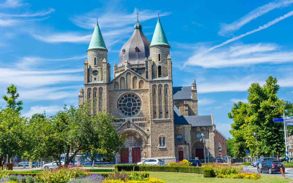 Romanische Kirche in Maastricht mit markanten Türmen, grünlichen Dächern und einer großen Rosette, umgeben von Bäumen, Blumen und einer gepflegten Parkanlage.