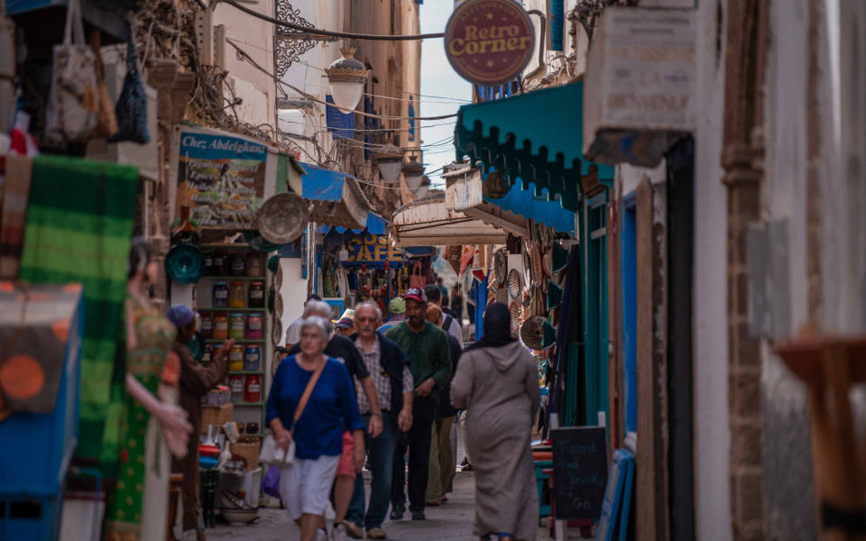 Belebte Gasse in der Altstadt von Essaouira mit Marktständen, Schildern, bunten Gewürzen und Touristen.