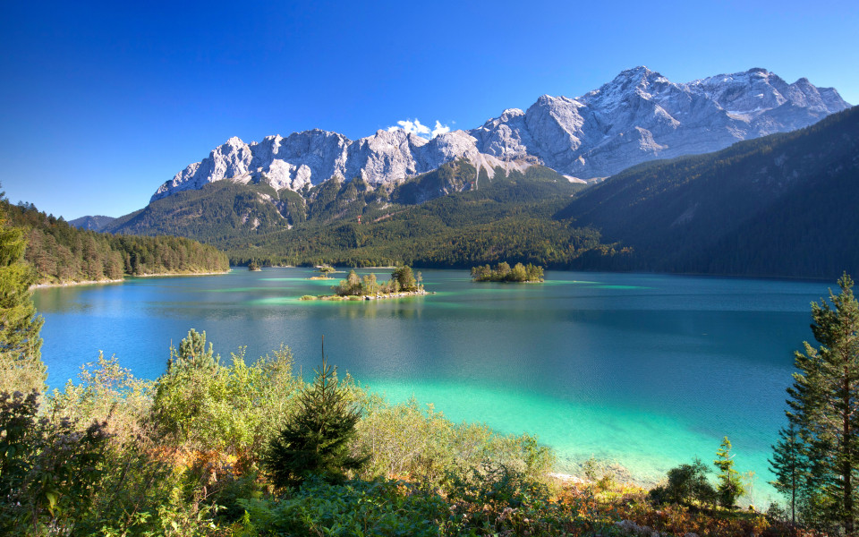 Blick auf den türkisfarbenen Eibsee mit kleinen Inseln, umgeben von dichtem Wald und den schneebedeckten Gipfeln der Zugspitze in Bayern, Deutschland. ChatGPT fragen