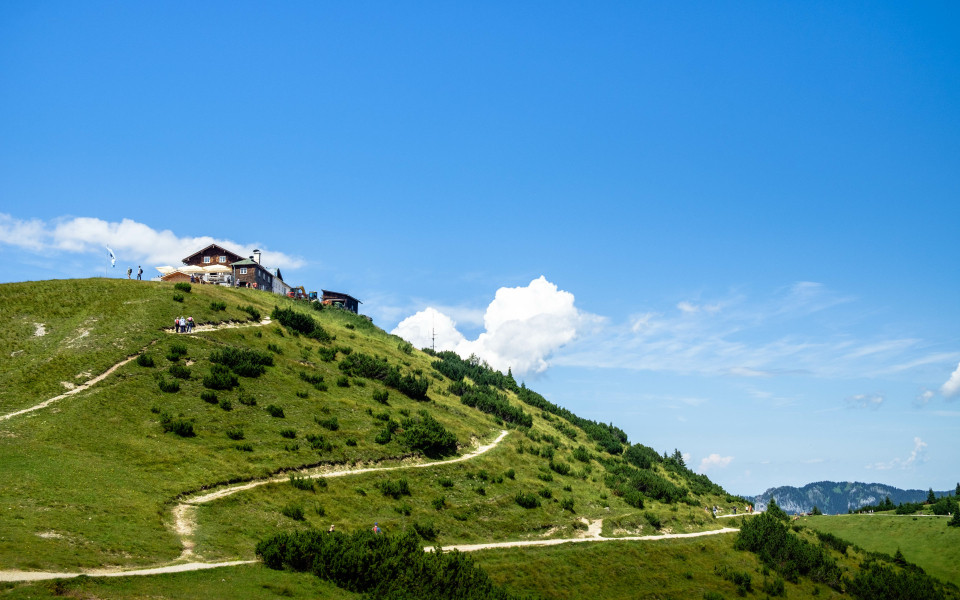 Wanderweg führt den grünen Hang des Wankbergs hinauf zu einer Berghütte, unter strahlend blauem Himmel mit wenigen Wolken.