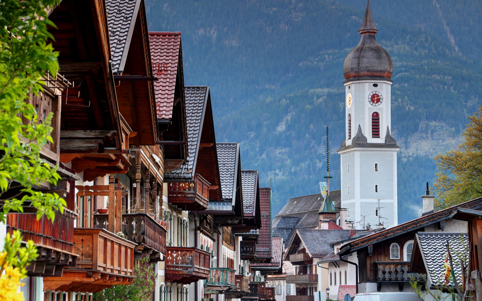 Die Ludwigsstraße in Partenkirchen mit traditionellen bayerischen Häusern und Balkonen und im Hintergrund der Zwiebelturm der Pfarrkirche St. Anton vor alpiner Bergkulisse.
